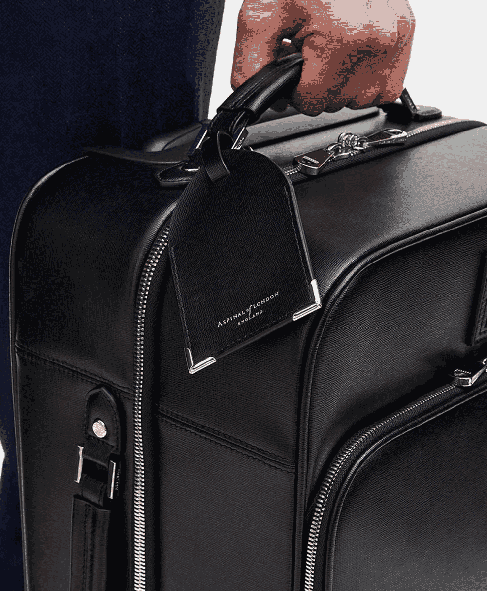 A close-up of a man in a dark suit holding a black leather Aspinal of London suitcase.