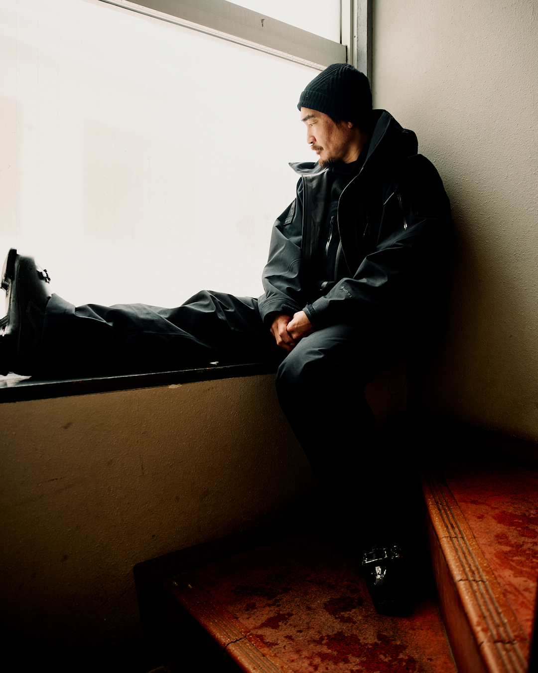 A man in all-black tactical gear sitting on a window ledge in a dimly lit, moody room.