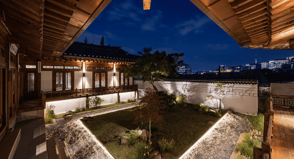 Night view of a traditional Korean hanok courtyard with illuminated pathways and a modern city skyline in the background.