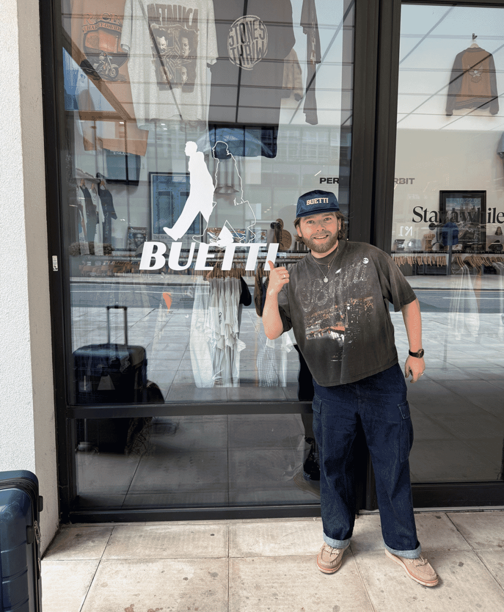 Man in a blue Buetti cap and graphic tee giving a thumbs up in front of a clothing store window.