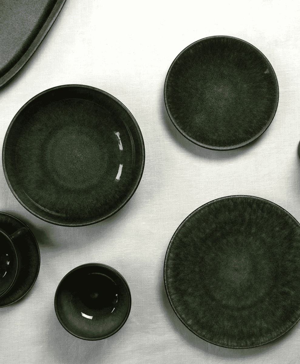 Flat lay of dark green ceramic plates and bowls on a white tablecloth.