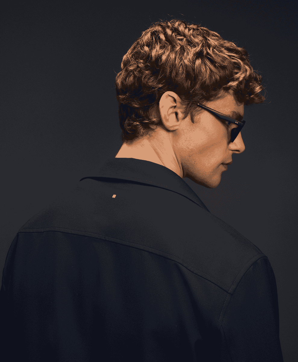 Profile shot of a man in a dark jacket with gold collar hardware and minimalist eyewear.