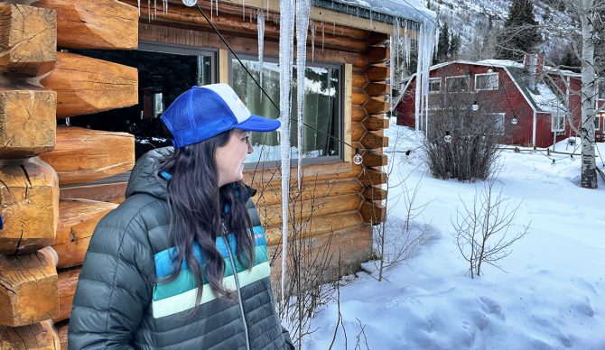 A woman wearing a striped Cotopaxi Fuego Down Parka standing near a wooden cabin.