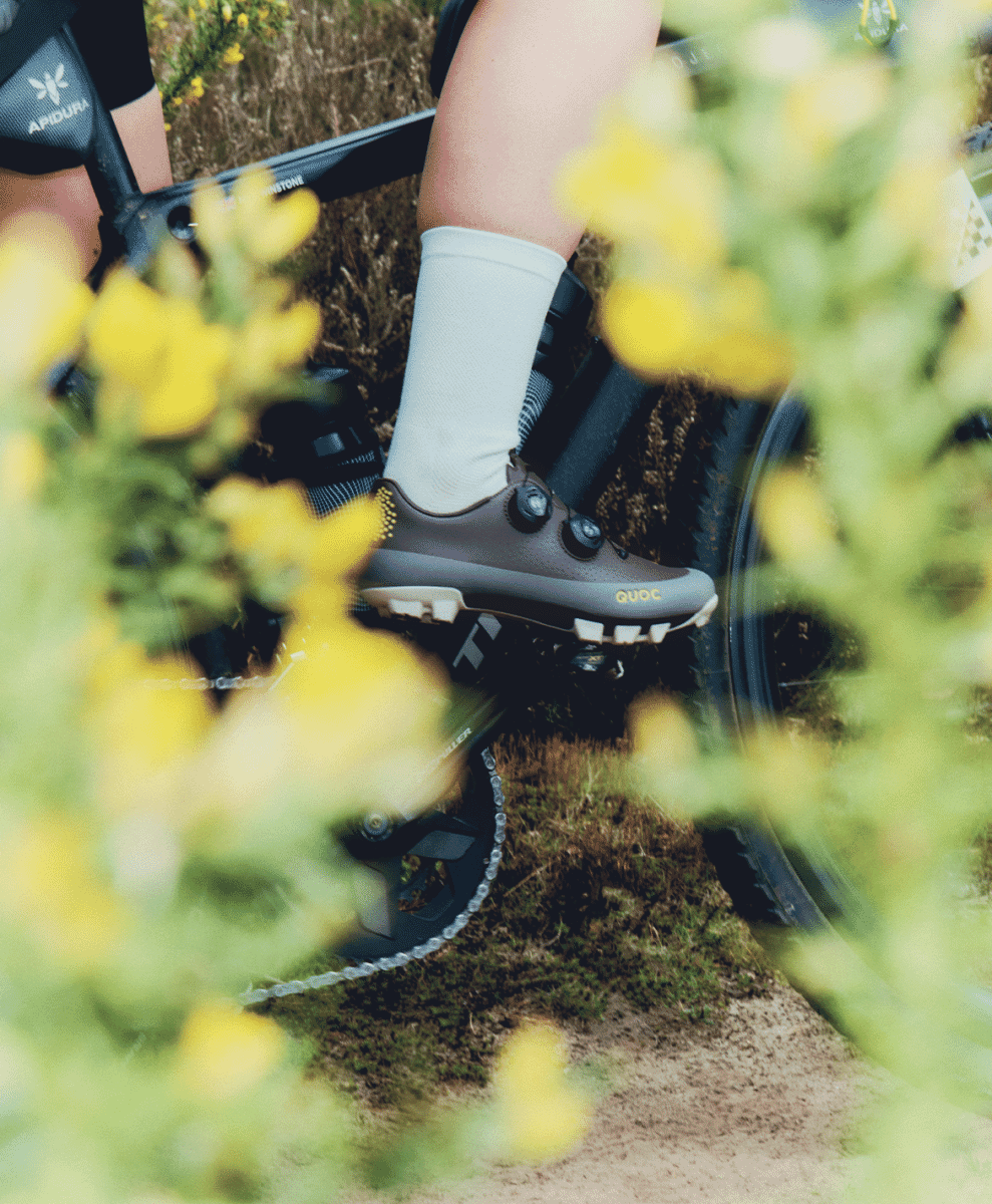 A cyclist wearing white socks and mountain biking shoes riding through a vibrant field of yellow flowers.