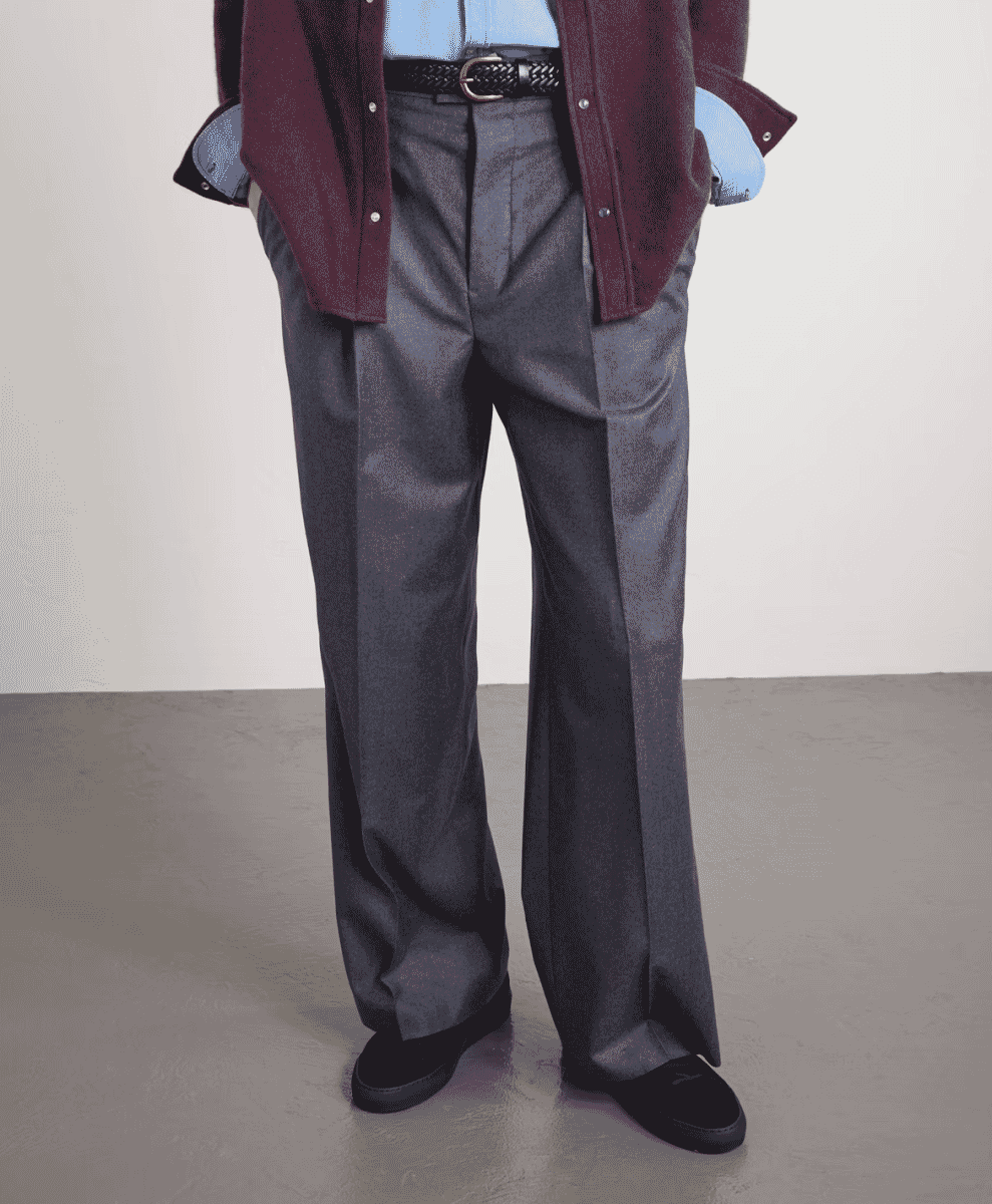 A man wearing grey pleated trousers and a maroon jacket in a studio setting.