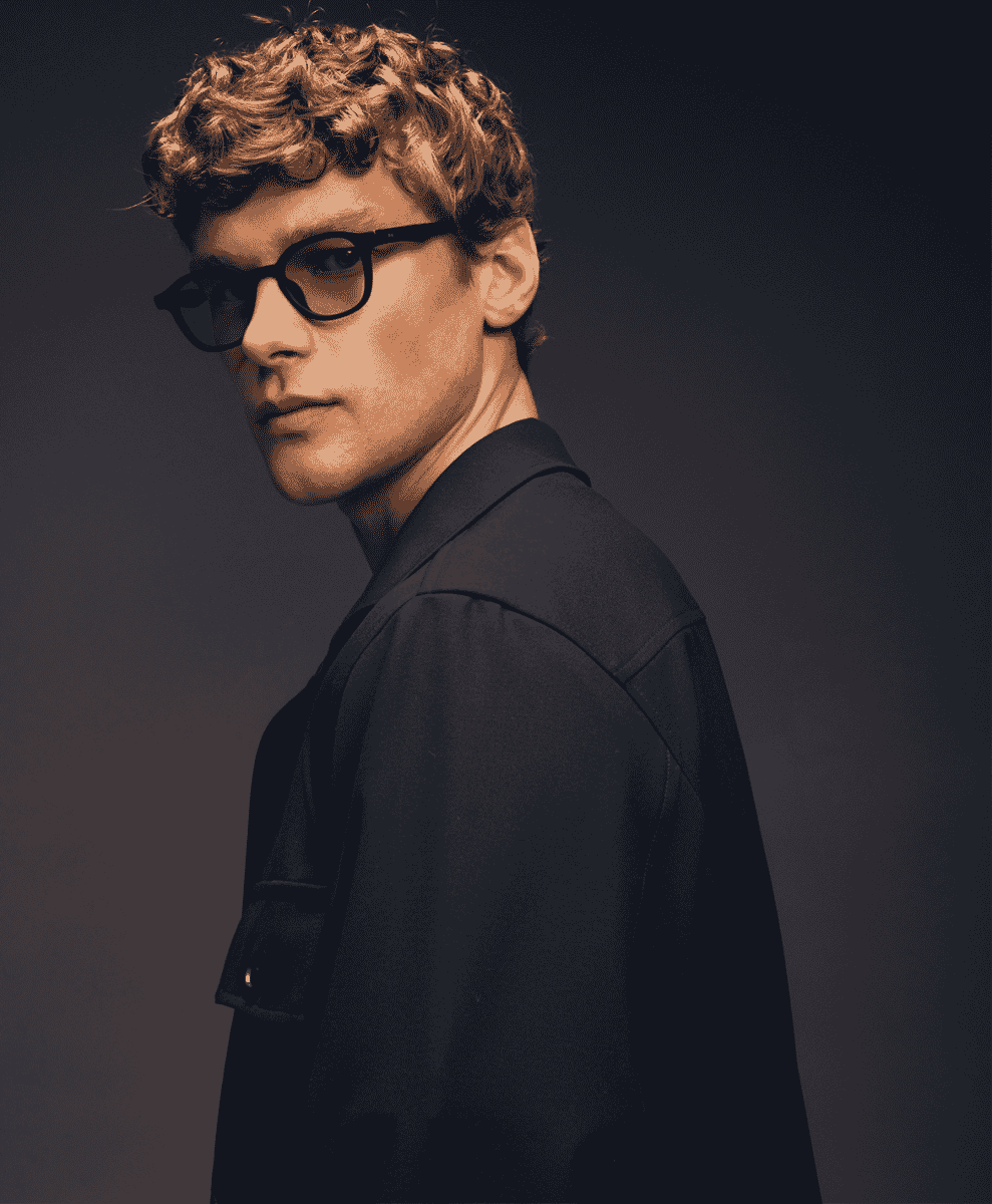 Man wearing black-rimmed glasses and a structured dark jacket over a collared shirt.