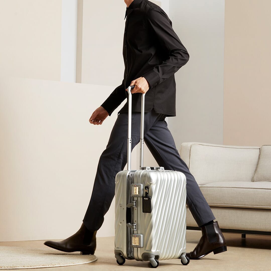 A man in business casual attire pulling a silver TUMI suitcase through a minimalist modern living room.