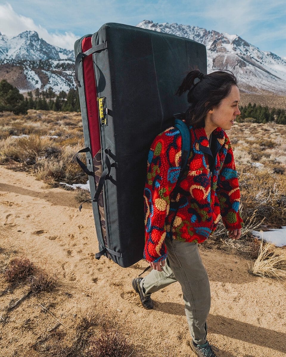A person carrying a climbing crash pad through a snowy landscape in a colorful fleece jacket.