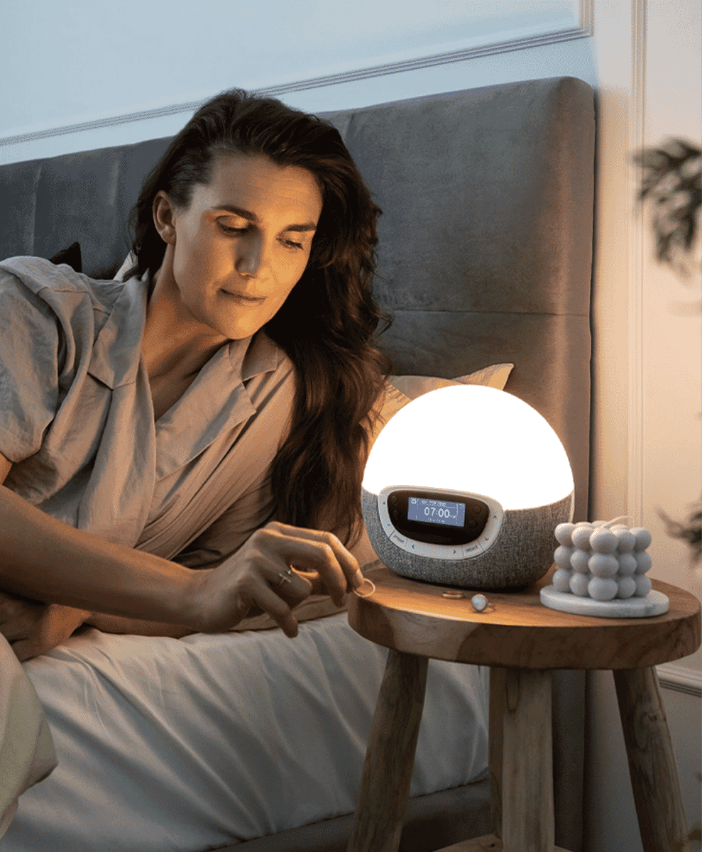 A woman in relaxed pajamas sitting in bed next to a glowing sunrise alarm clock on a wooden nightstand.