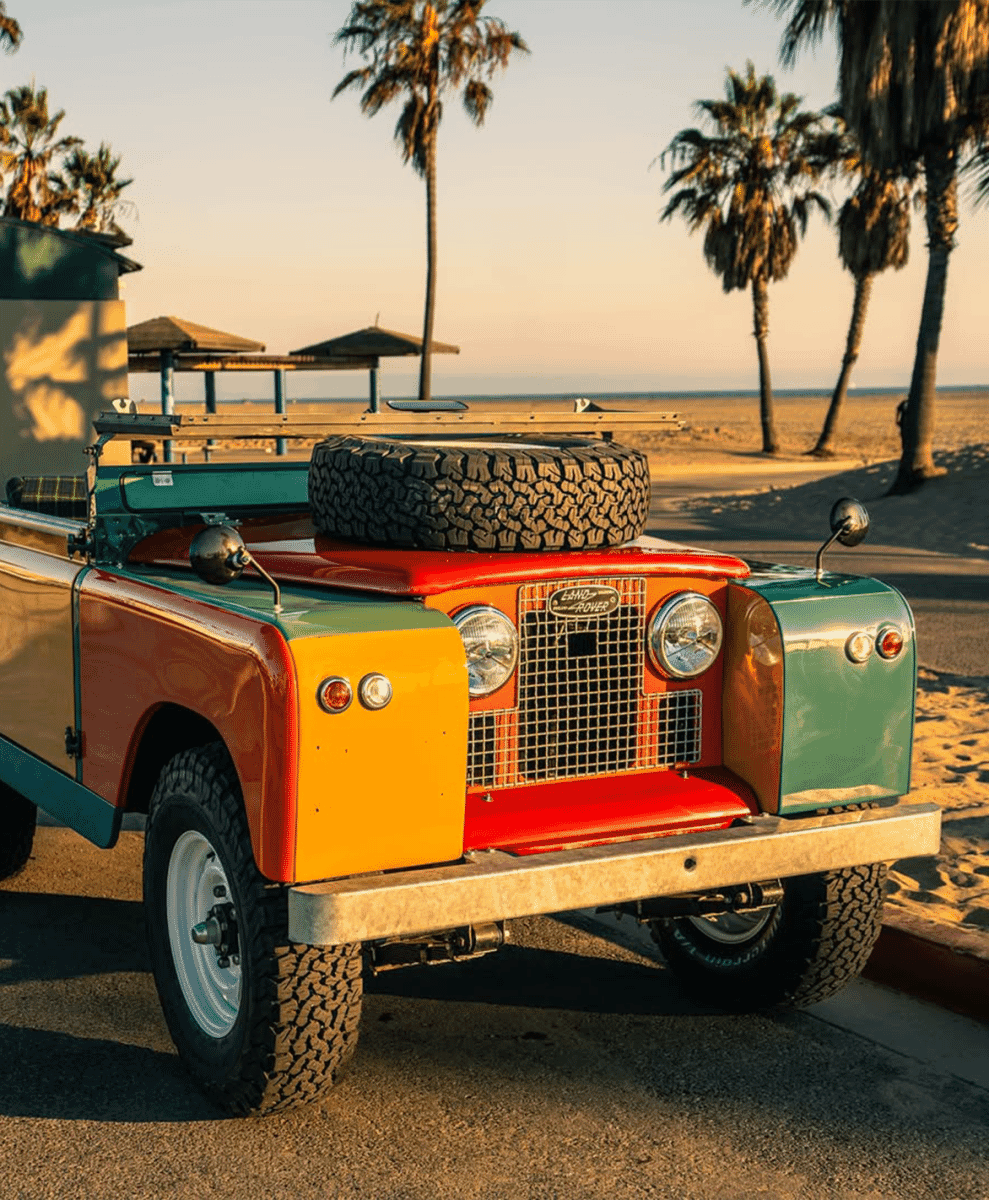 A multicolored Land Rover with a spare tire on the roof parked on a coastal street at sunset.