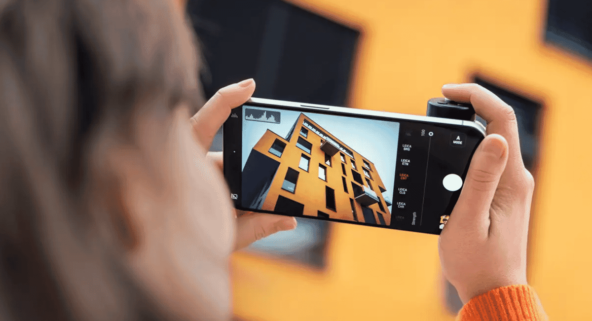 A person in an orange sweater using a smartphone to capture a modern yellow building.
