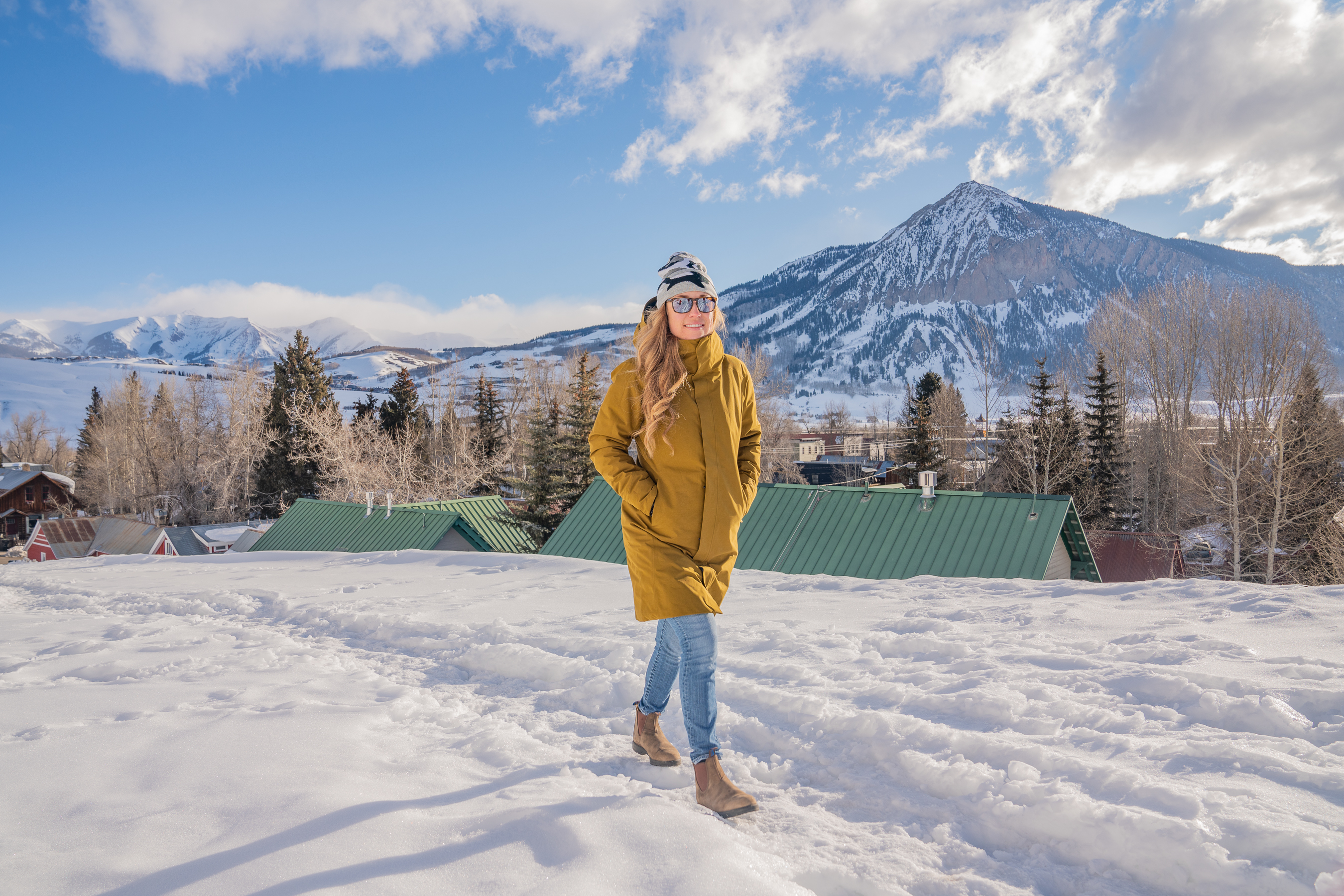 A person in a black winter parka walking on a snow-covered trail with mountains in the background.