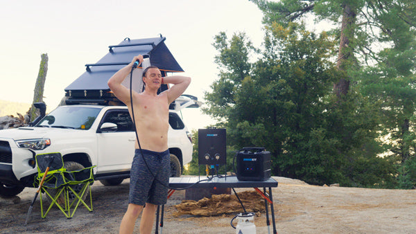 A portable camping shower system placed among outdoor gear in a natural campsite setting.