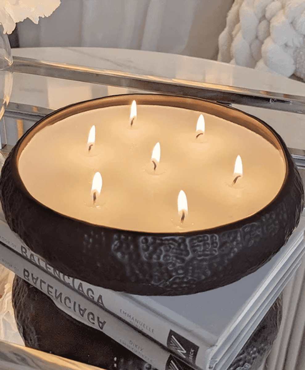 A lit six-wick candle in a textured black bowl resting on a stack of books.