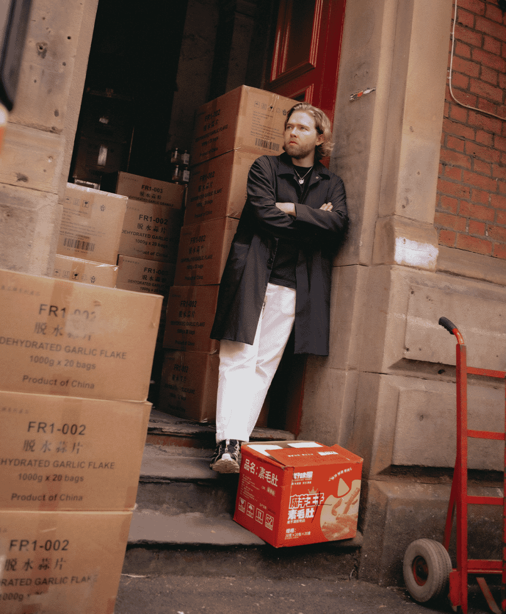Full body shot of a man in a black coat and white wide-leg pants in an urban alleyway.