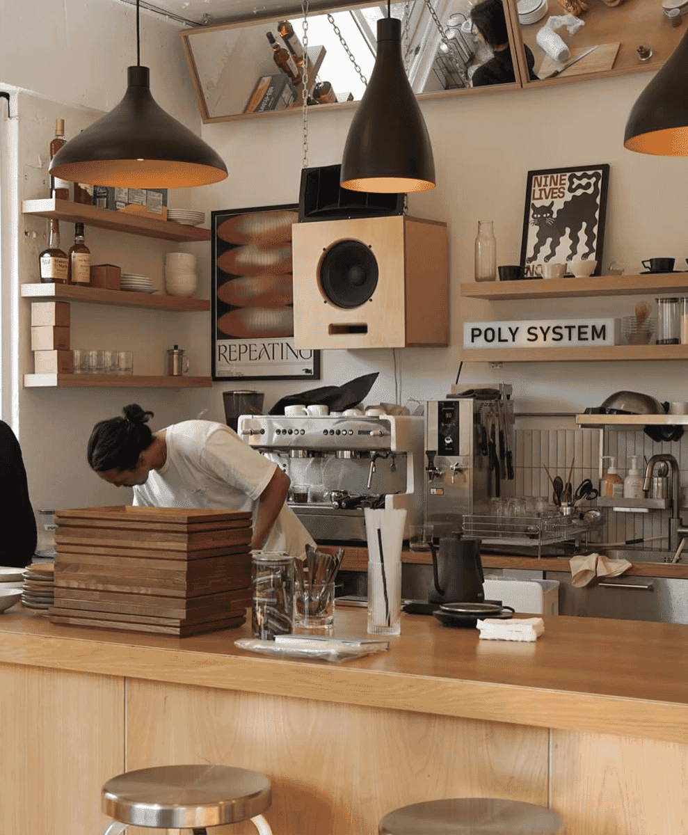 A barista in a white t-shirt working behind a wooden counter in a modern, minimalist café under pendant lights.