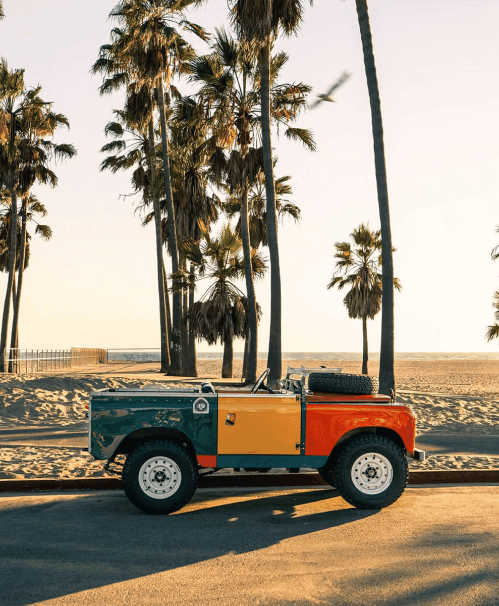 A vibrant, multi-colored vintage Land Rover convertible parked on a sunny coastal road with palm trees.