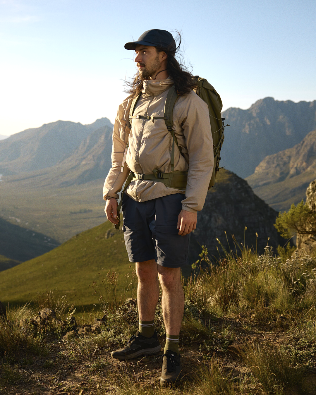 Mid-shot of a hiker wearing a Fjällräven beige windbreaker against a backdrop of sunlit mountain peaks.