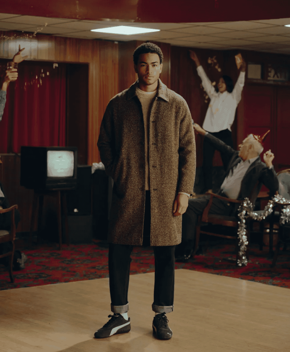 A man wearing a brown textured wool coat over a cream sweater in a retro-style party room.
