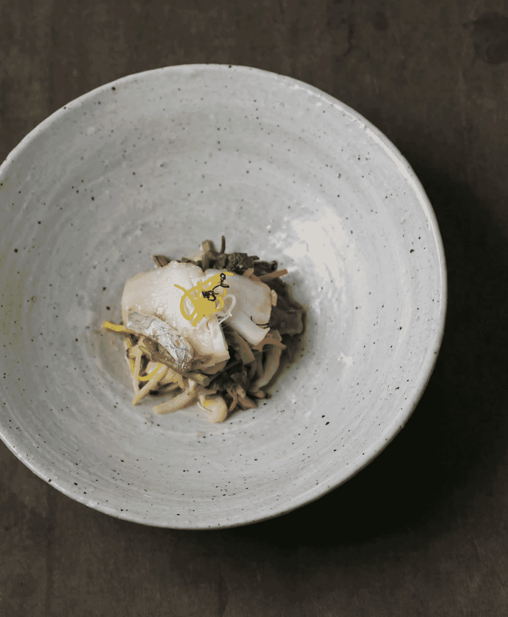A minimalist ceramic bowl holding a delicately plated dish of fish and mushrooms on a dark wooden surface.