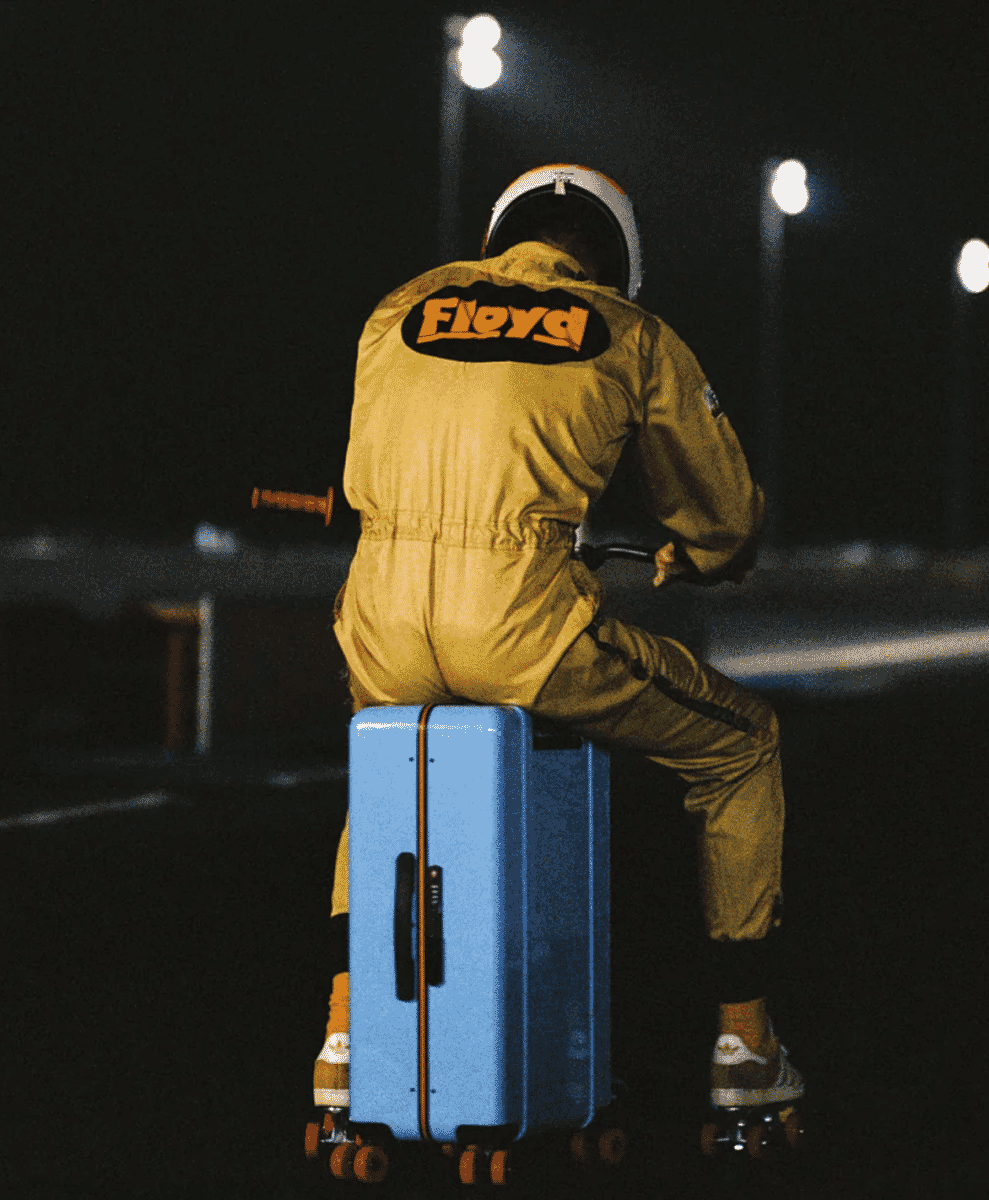 A person in a yellow Floyd racing jumpsuit riding a blue suitcase on roller skates at night.