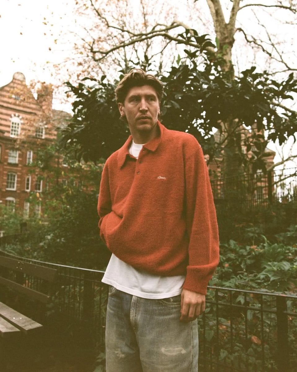 Man wearing a textured orange knit polo sweater in an urban park setting.