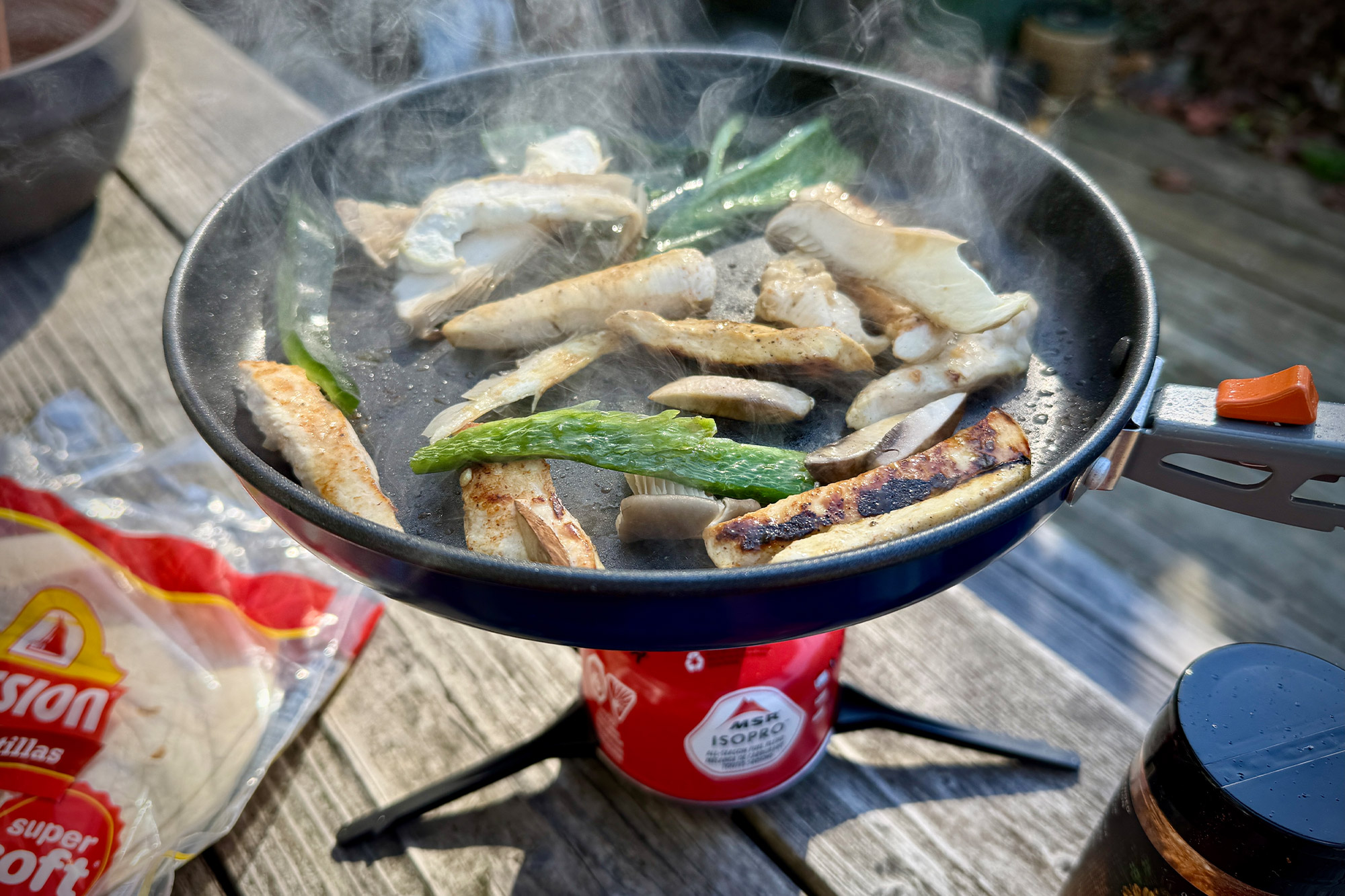 Sizzling chicken and peppers being sautéed in a frying pan over a camping stove.