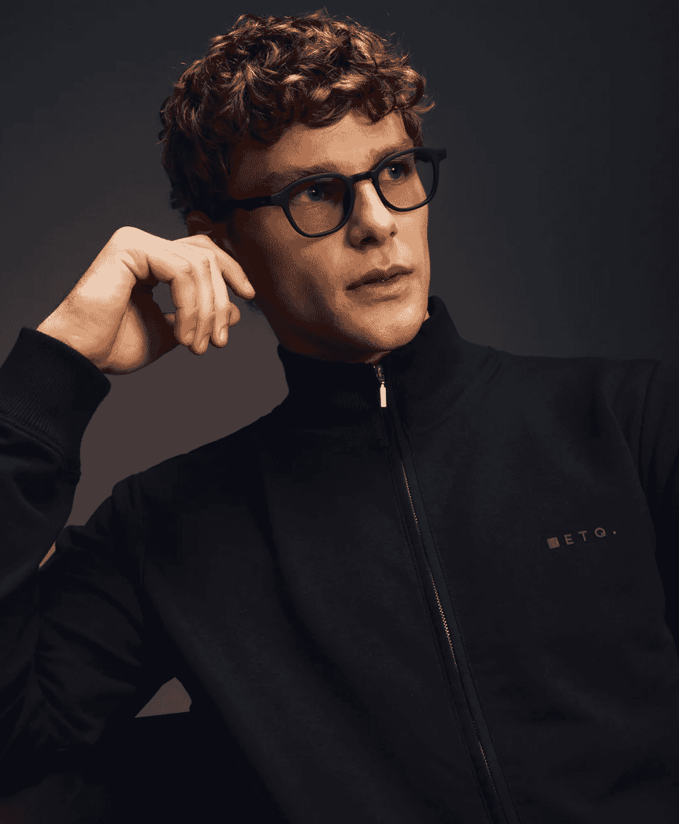 Man in an ETQ branded black zip-up jacket and black-rimmed glasses.