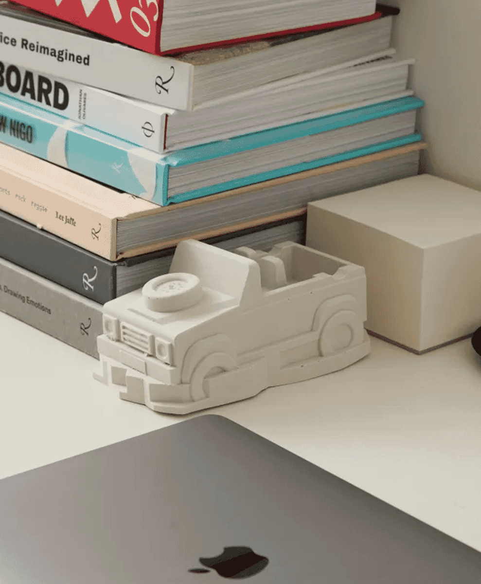 A white concrete model of an off-road vehicle on a desk next to books and a laptop.