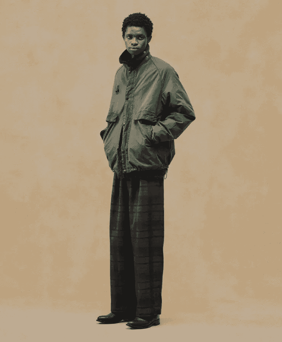 Full-length studio portrait of a man wearing an olive-green cropped jacket and wide-leg plaid trousers.