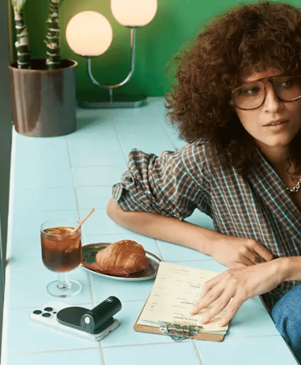 A woman in a plaid shirt at a café table with a coffee, croissant, and a notebook.