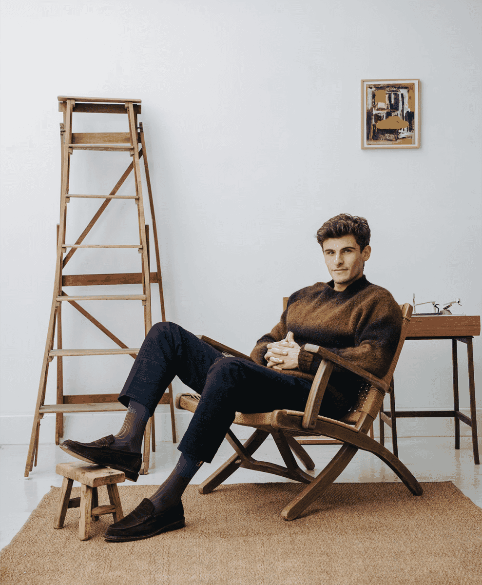 A man in a brown textured sweater and dark trousers sits relaxed in a minimalist studio setting.