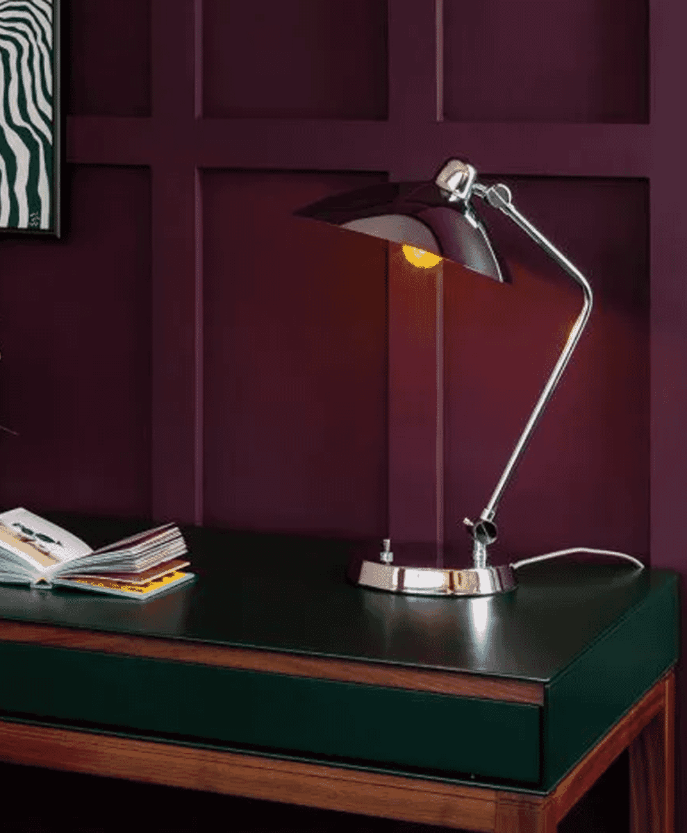 A modern chrome-finished desk lamp illuminating a dark green leather-topped desk against a burgundy wall.