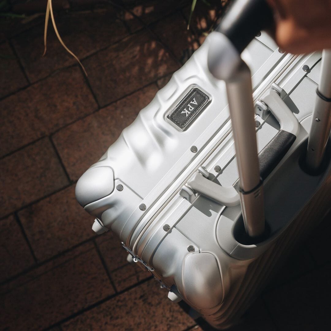 Close-up of a silver grooved suitcase with a customized APK monogram plate on a brick surface.