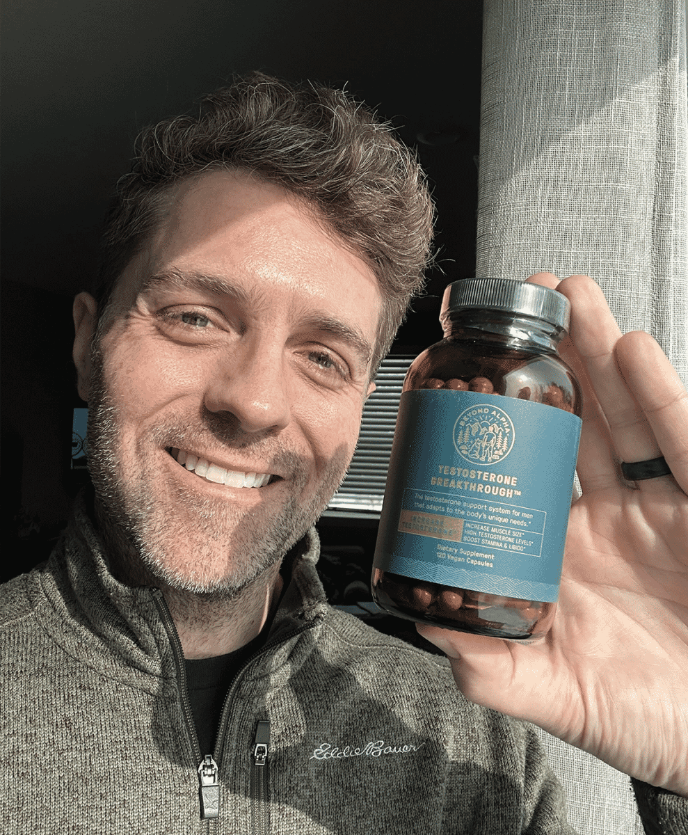 A man in a grey Eddie Bauer quarter-zip sweater smiling while holding a supplement bottle.