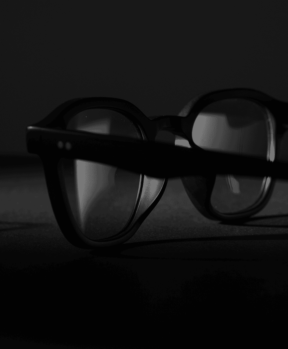 Monochrome still life of black-framed rounded eyeglasses on a dark surface.