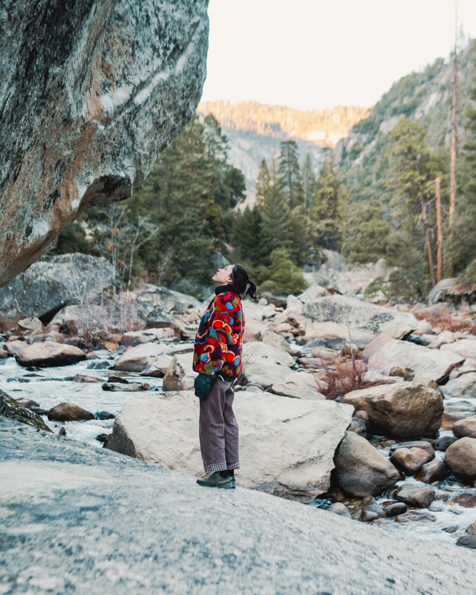 A woman in a colorful patterned knit sweater standing by a mountain stream.