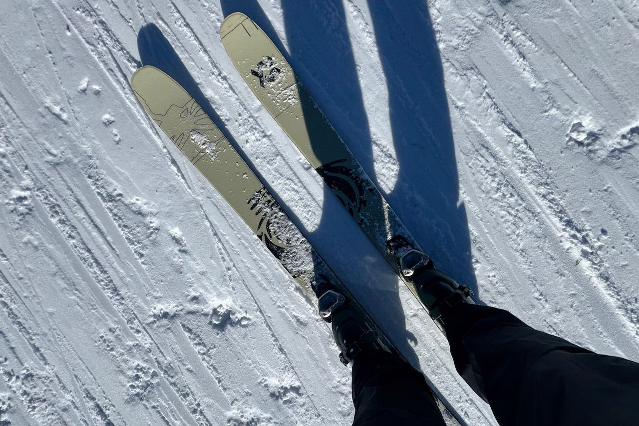 Point-of-view shot looking down at the tips of Völkl Revolt 101 skis while standing on a snowy mountain slope.