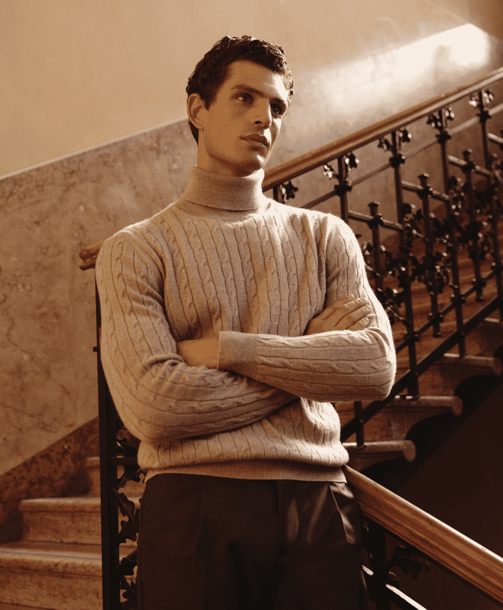 A man in a cream-colored cable-knit turtleneck standing on an ornate staircase.