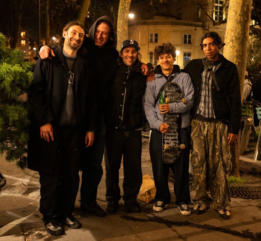 A group of five men in streetwear posing on a Parisian sidewalk at night with a skateboard.