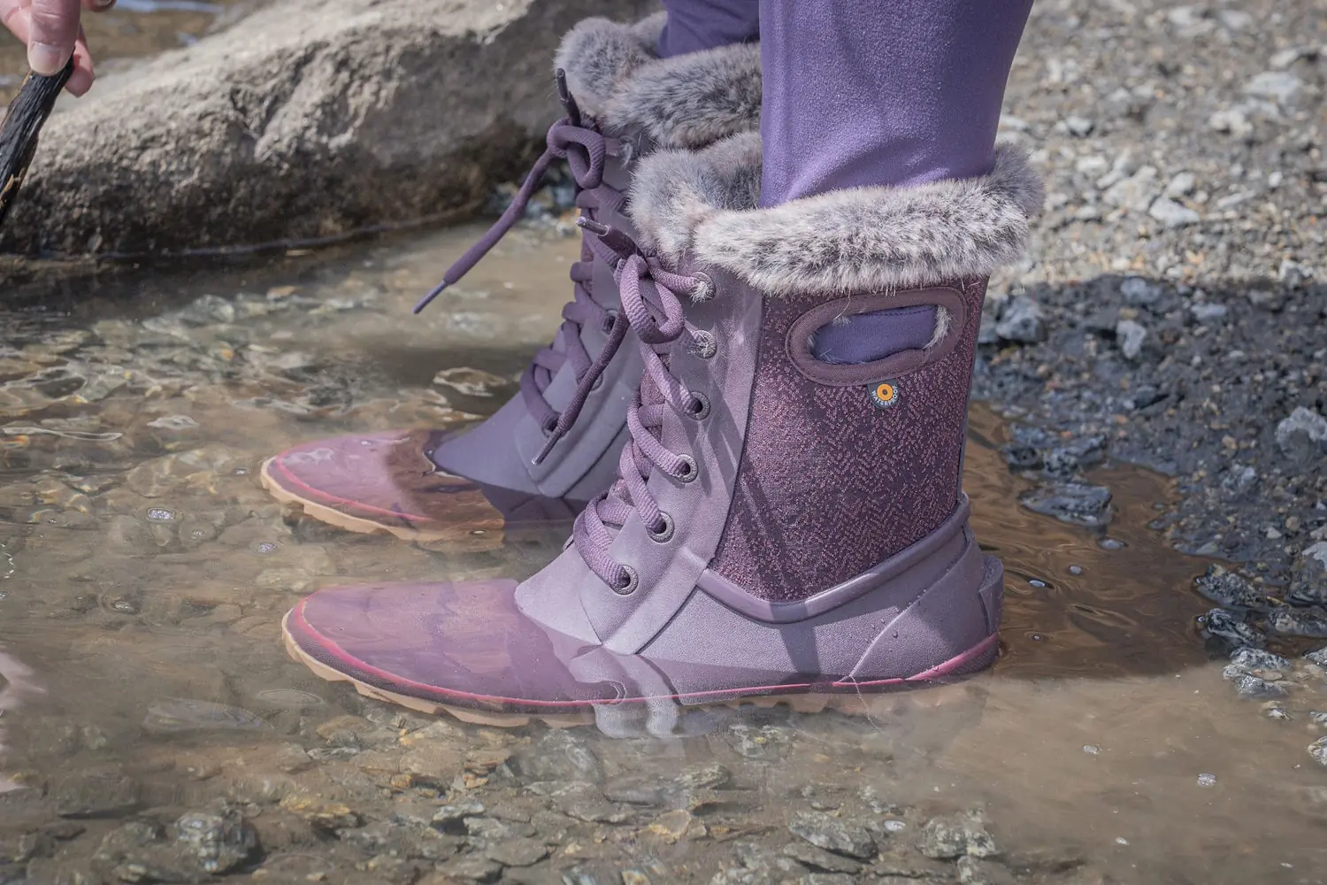 A pair of Bogs Arcata Faded boots submerged in a shallow, rocky puddle of water.