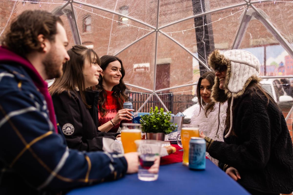 A group of friends laughing and drinking inside a transparent heated winter igloo.