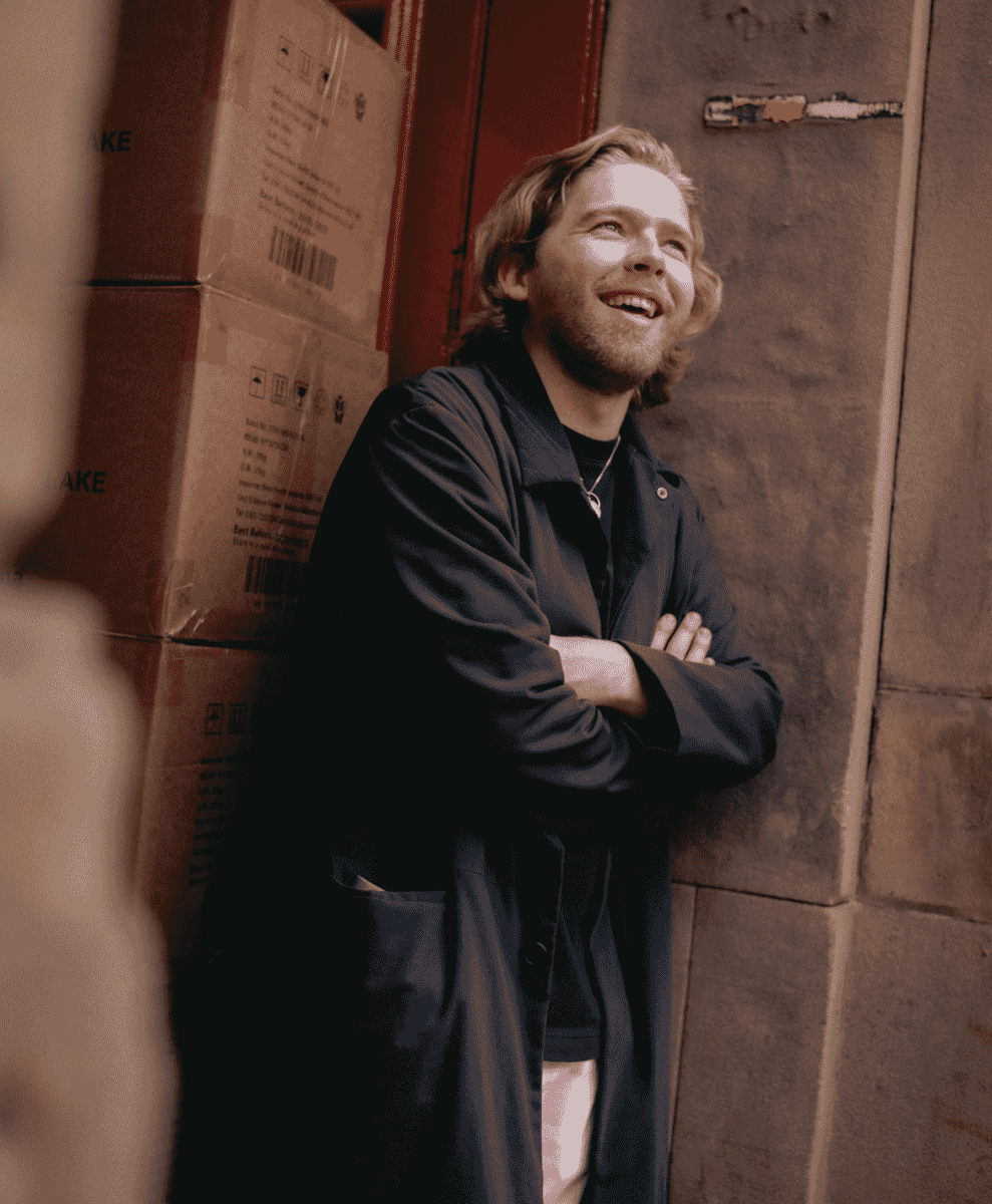 Man in a long dark trench coat smiling while leaning against a brick wall with cardboard boxes.