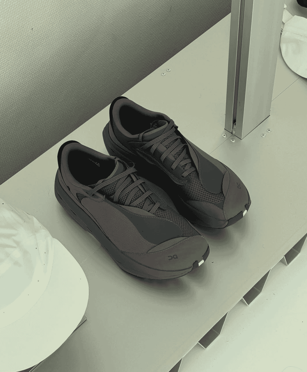 Dark gray ON running shoes on a minimalist white shelf next to caps.