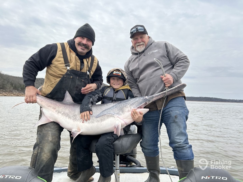 A group of three anglers on a boat smiling and holding a trophy-sized paddlefish.