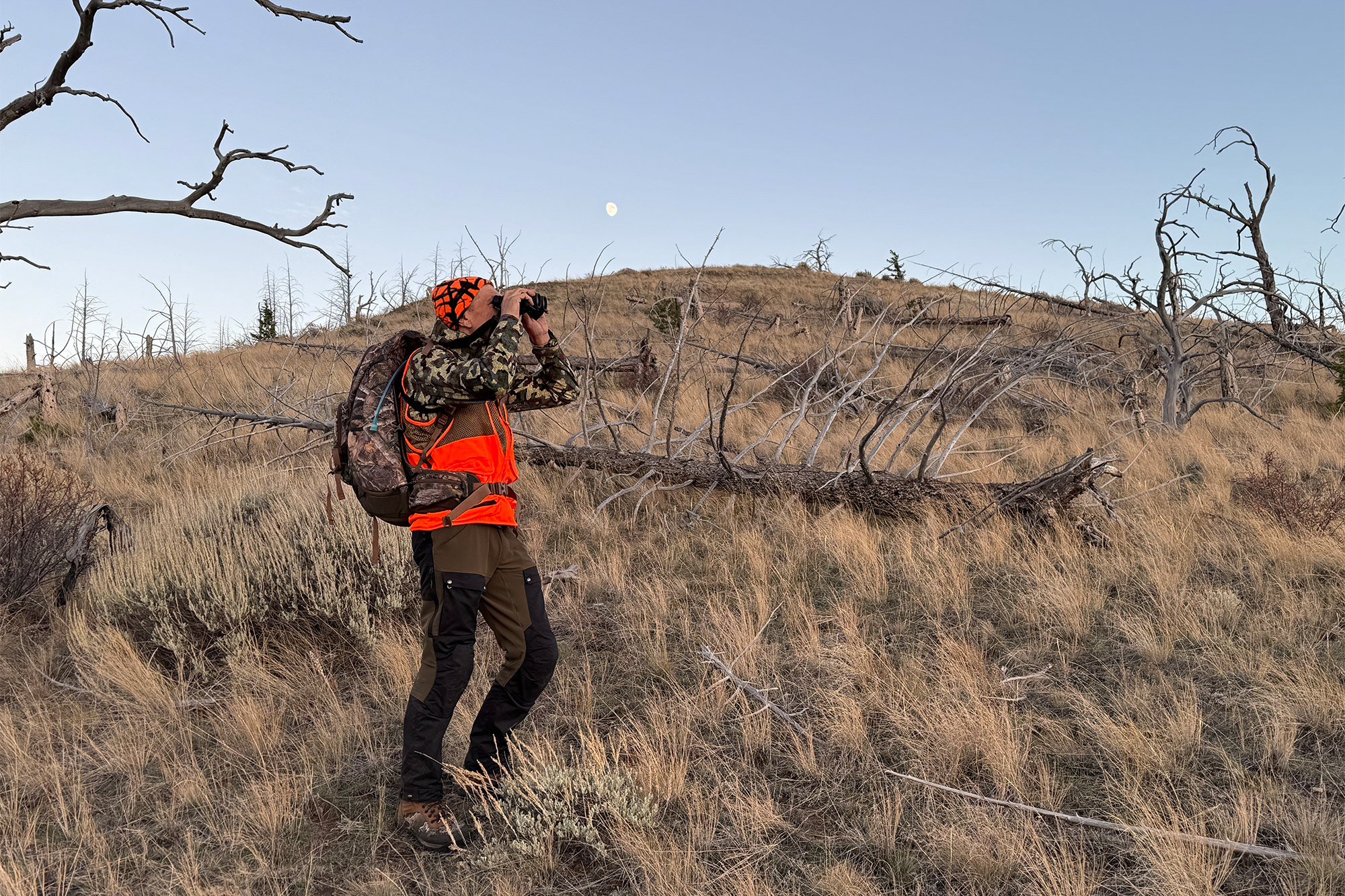 A hunter standing on a ridge glassing a distant hillside with binoculars.