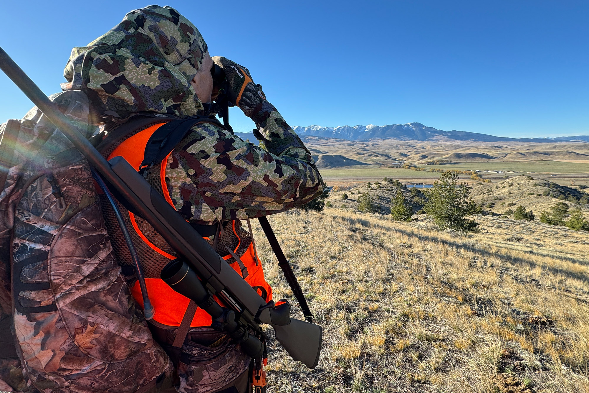 A hunter in a camo soft shell jacket using binoculars on a grassy hillside.