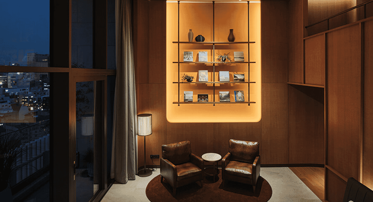 A warmly lit interior lounge with leather armchairs and a glowing bookshelf, overlooking a city skyline at night.