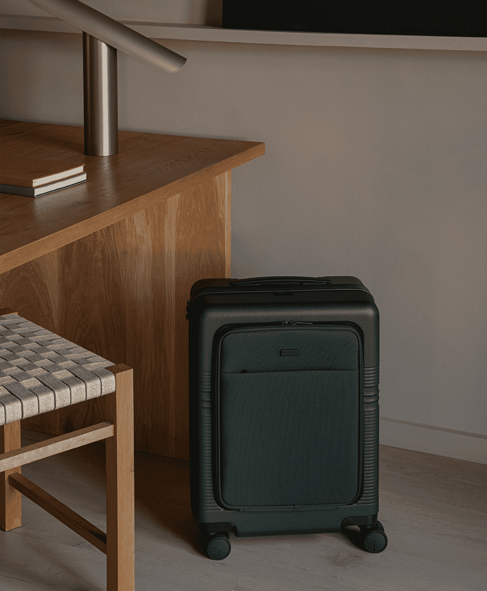 A black NORTVI front-pocket carry-on suitcase placed in a minimalist room beside a wooden desk.