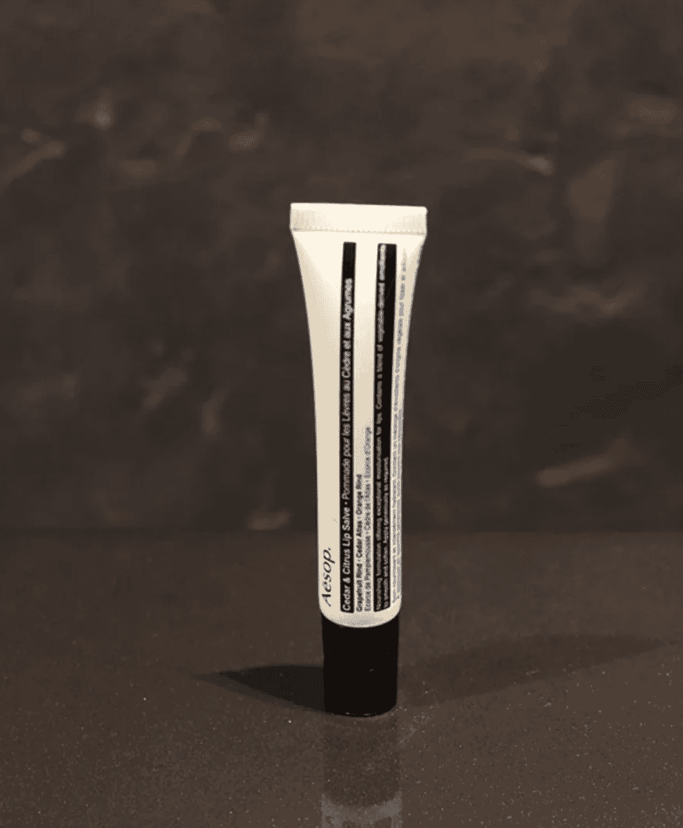A tube of Aesop Cedar & Citrus Lip Salve standing on a dark reflective surface.